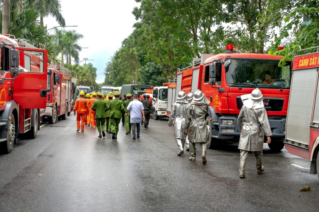 Firefighters in uniforms and fire trucks responding to an emergency on a city street with lush greenery.
