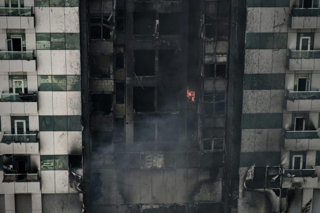 A high-rise building's facade shows severe fire damage with lingering smoke.