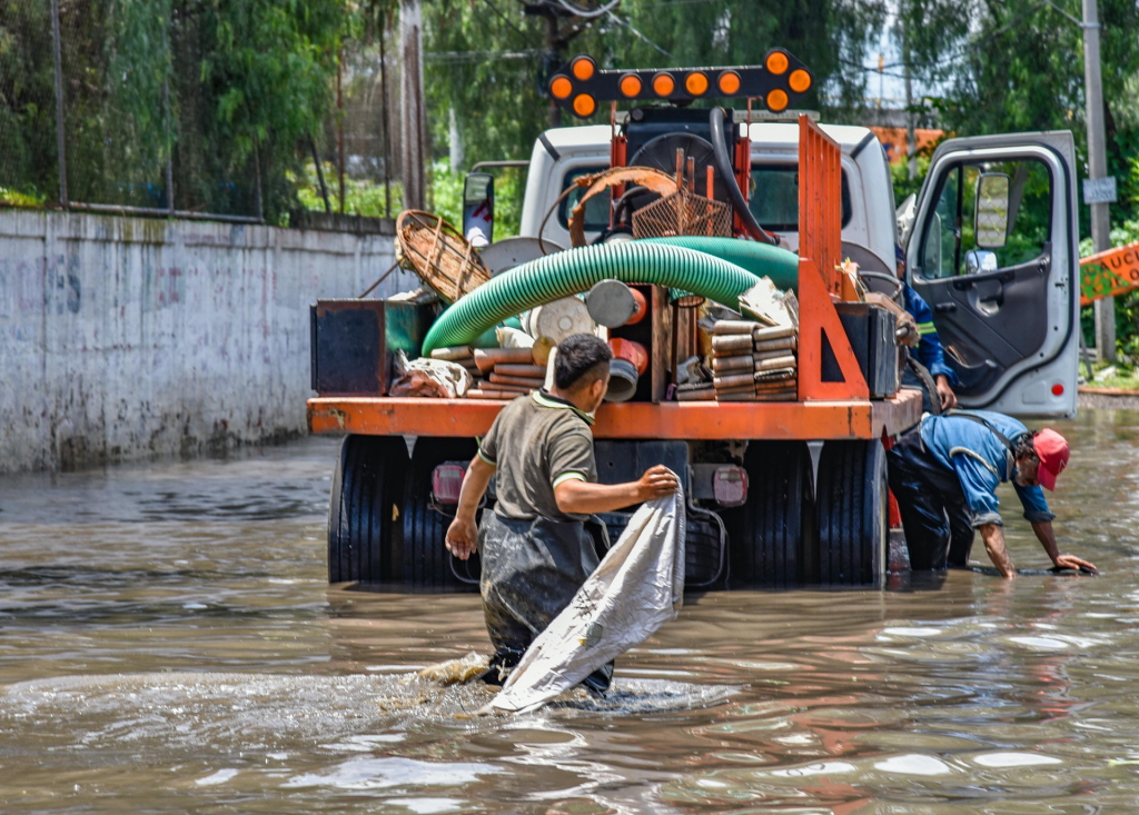 Workers wading through floodwaters near a truck, managing a city crisis after heavy rain.