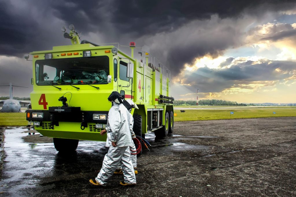 Firefighters in protective gear near a fire truck on a runway with dark storm clouds.