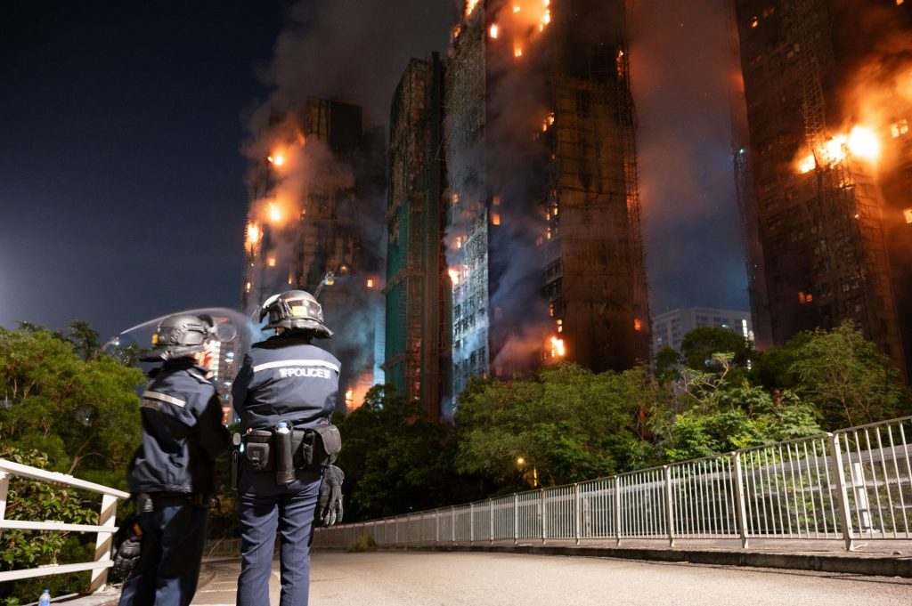 Riot police watch buildings engulfed in flames at night.